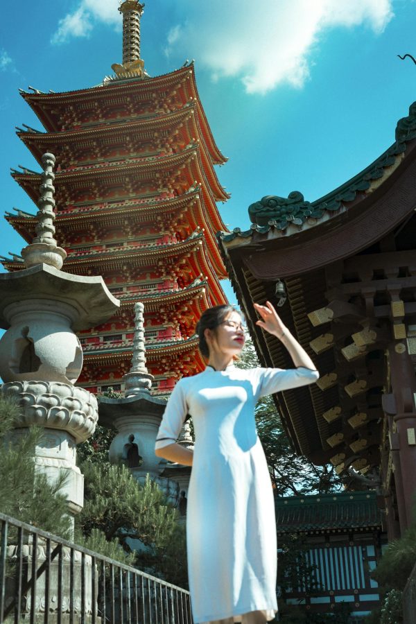 From below of beautiful young Japanese female in traditional white dress covering face by hand from bright sunlight while standing against amazing background of Sensoji temple ancient sculptures and cloudy blue sky